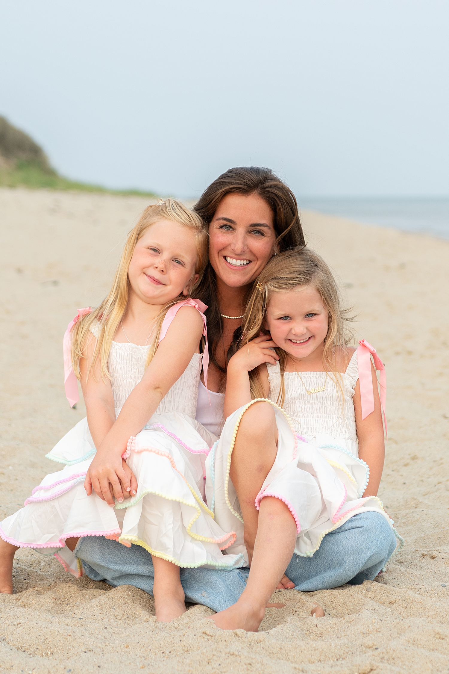 brunette sits on sand with two daughters in her lap after learning when to book your Nantucket family photos
