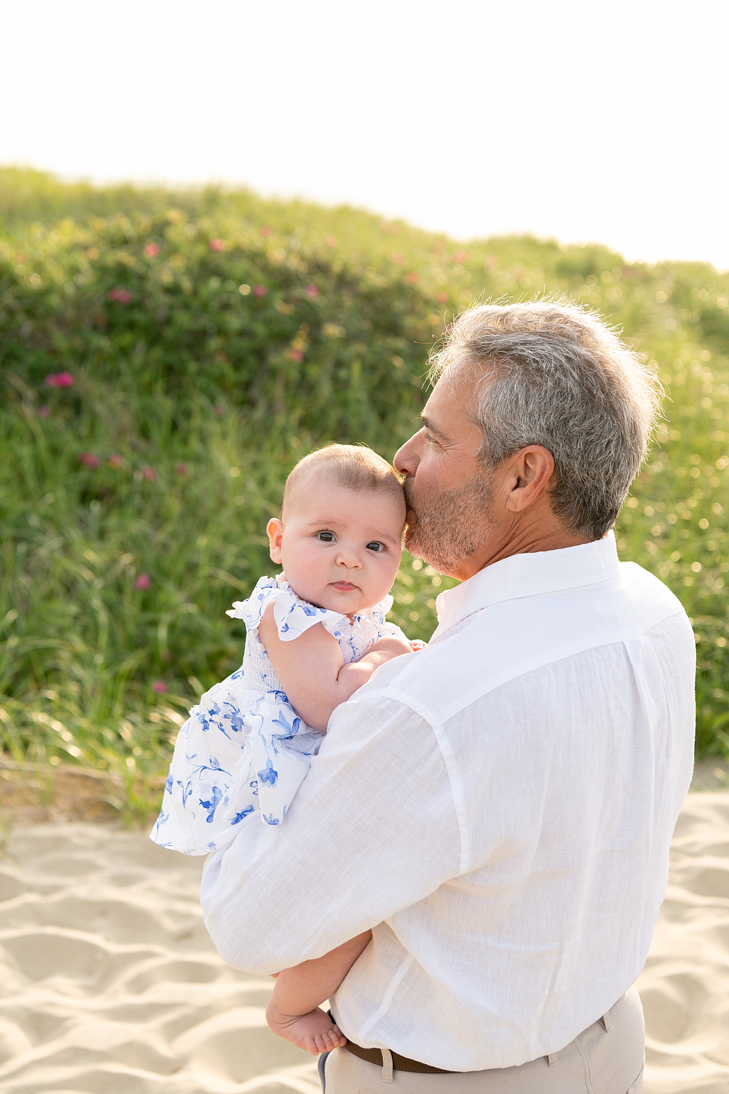 grandfather holds little one and kisses her head while standing in the sand by Nantucket photographer