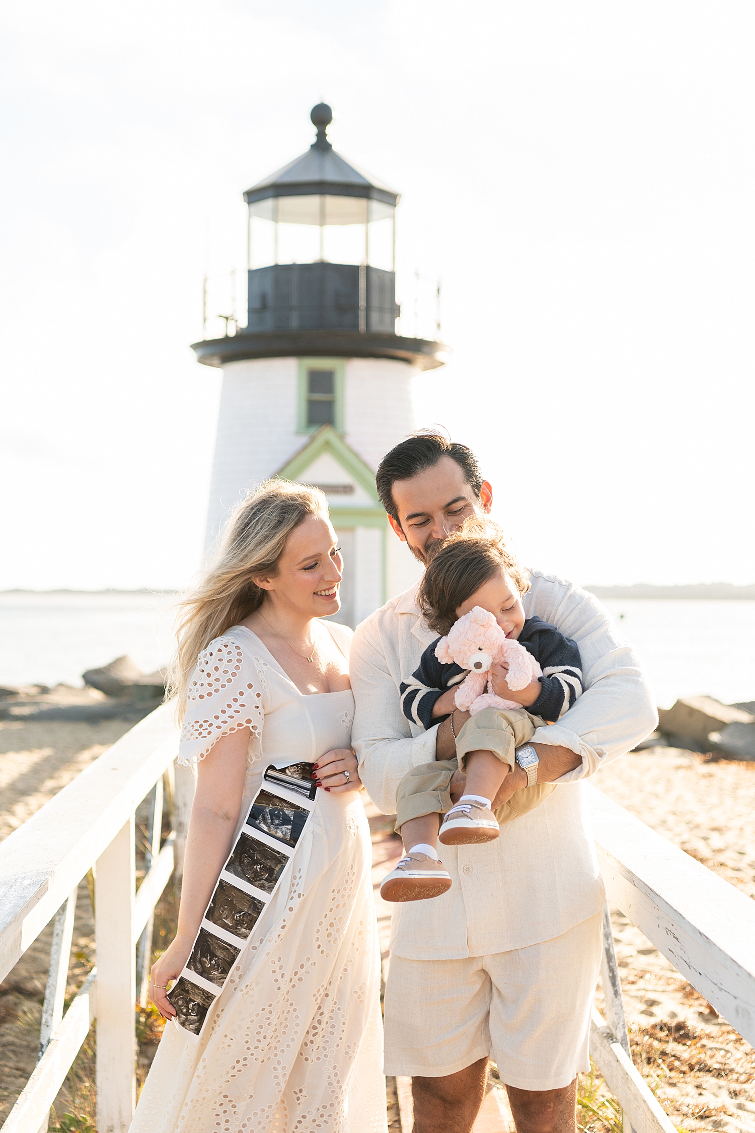 family snuggles in front of a lighthouse, showing off sonogram photos by Nantucket photographer