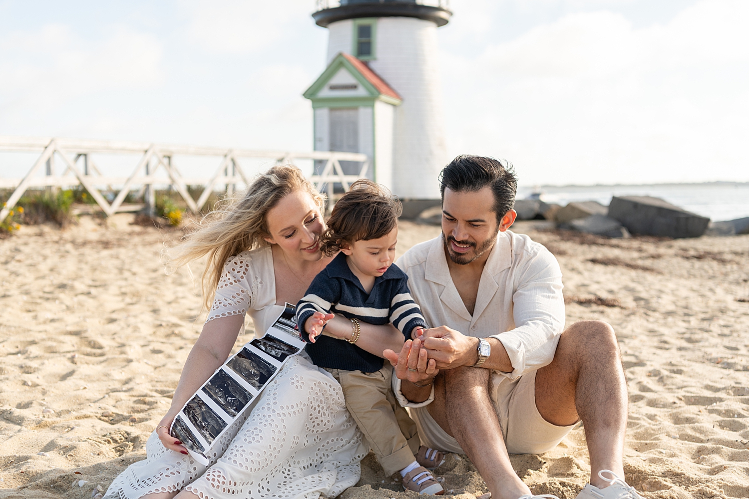 mom and dad sit in front of lighthouse with toddler and sonogram photos by Nantucket family photographer