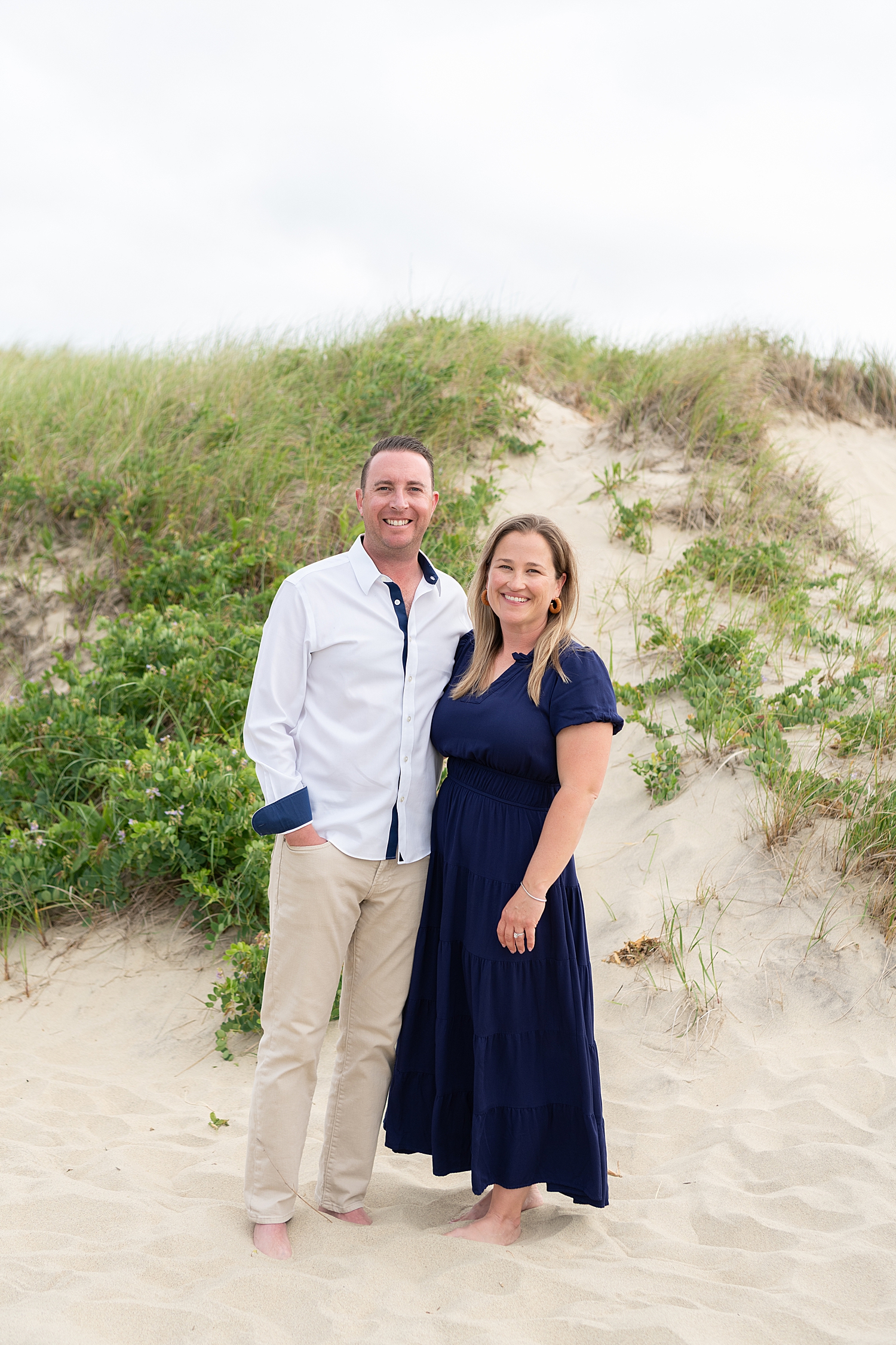 mom and dad stand next to sand dune by Nantucket Family Photographer