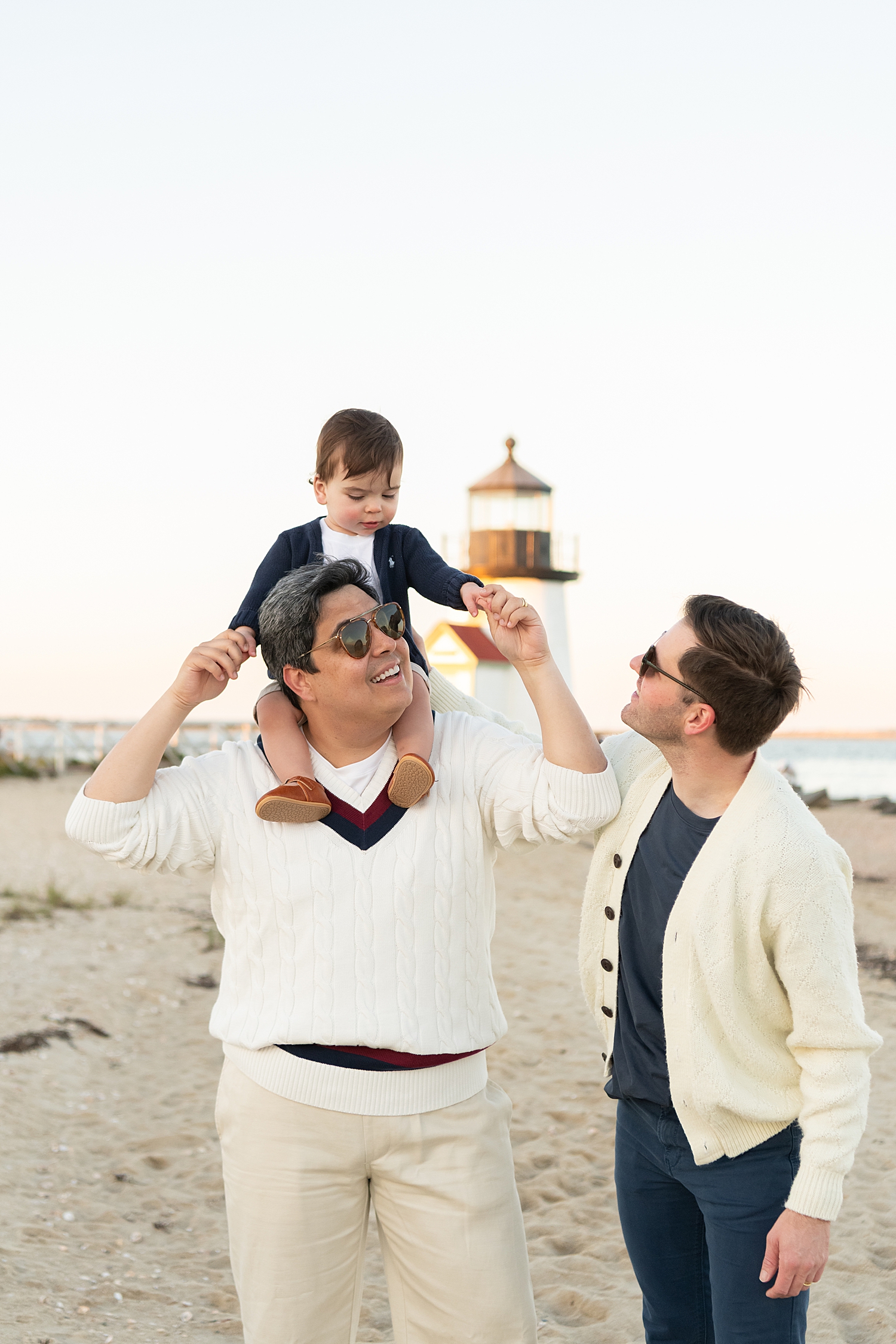 little one sits on dad's shoulder next to other dad looking up for 1st birthday photos with grandparents
